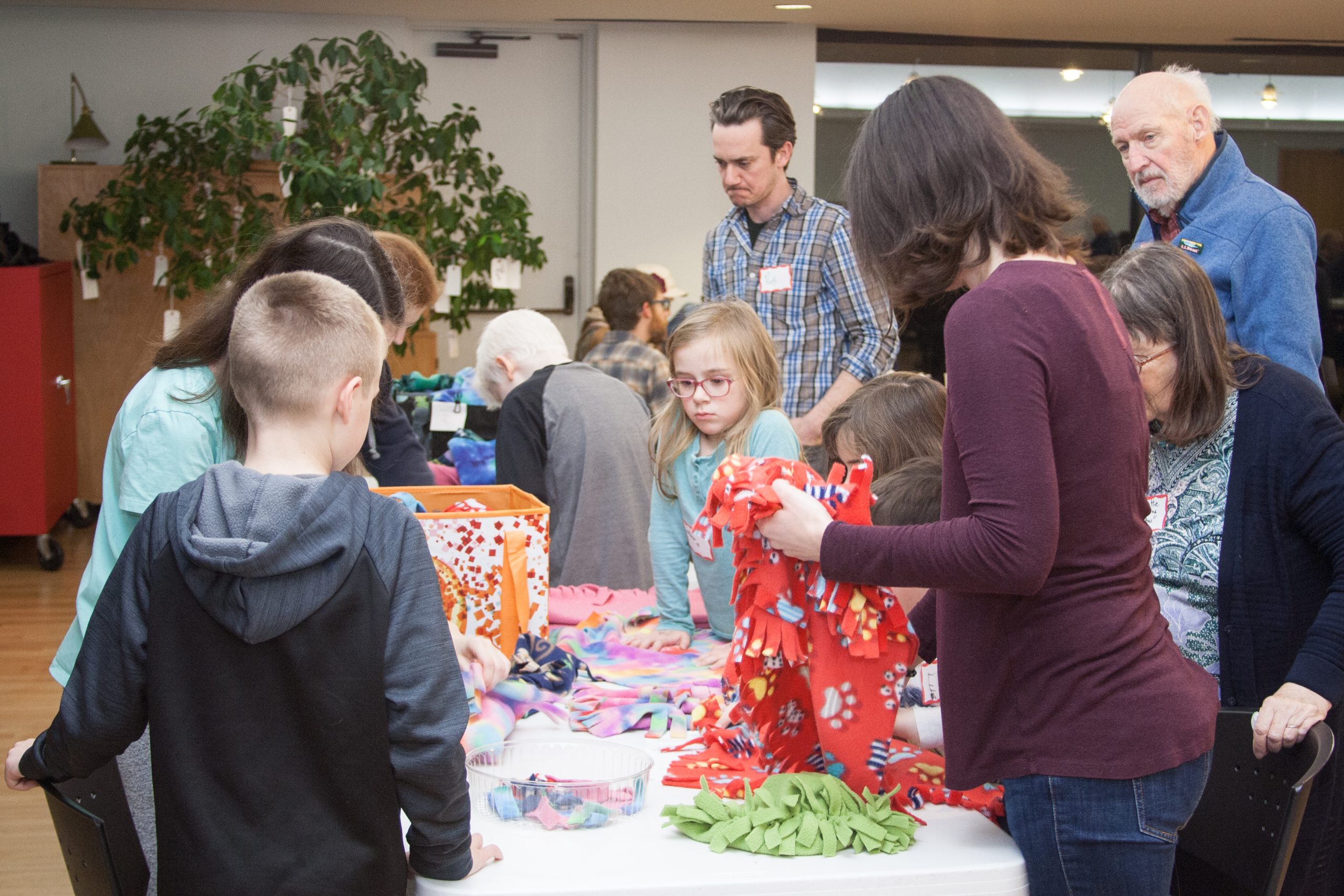 Several children and adults gather around a table learning how to make fleece hats.